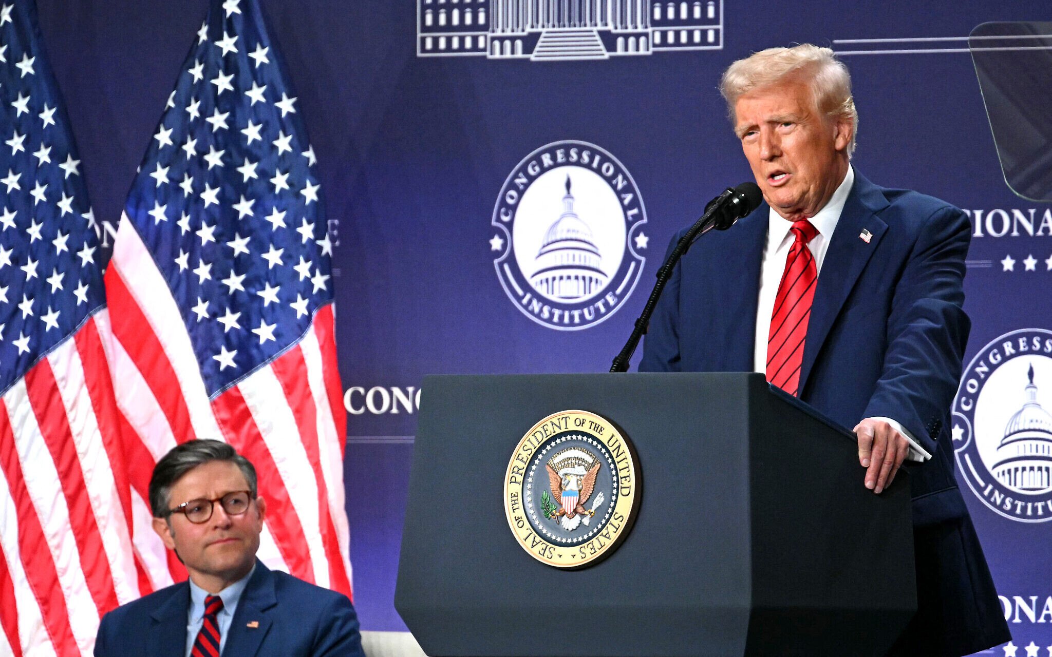 A U.S. political leader speaking at a podium with the presidential seal, flanked by American flags, during a formal announcement.